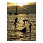 Silhouetted boats, and figures of children at play, against the setting sun reflected in the water.